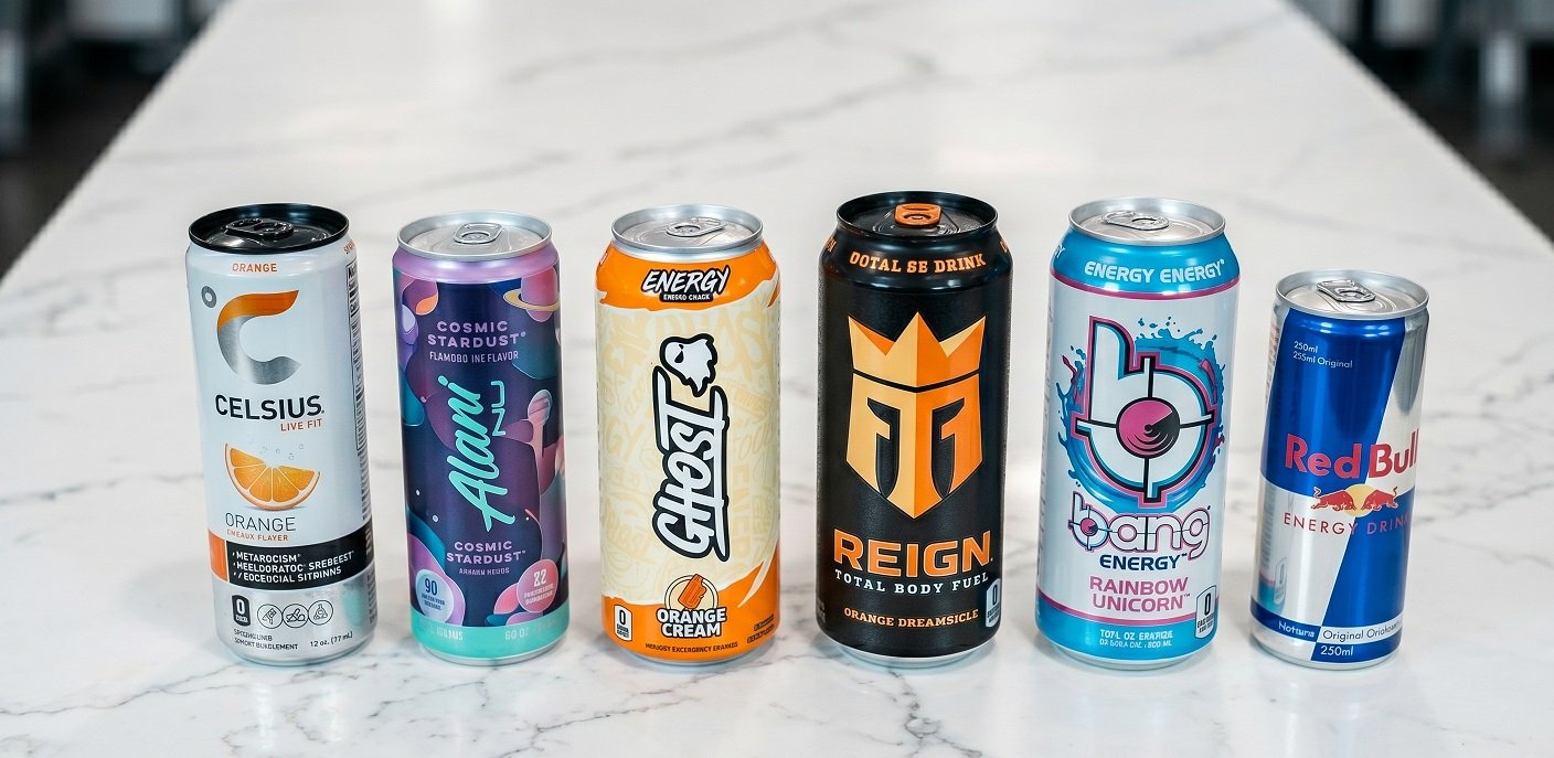 A lineup of energy drink cans including Celsius, Alani Nu, Ghost, Reign, and Red Bull arranged on a white surface for comparison