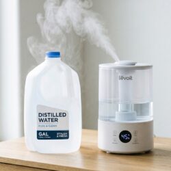 Gallon jug of distilled water next to a white ultrasonic humidifier on a wooden surface with clean mist rising