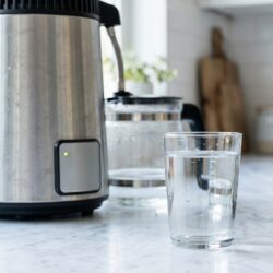 Glass of distilled water beside a countertop water distiller on a clean kitchen counter, illustrating what distilled water is